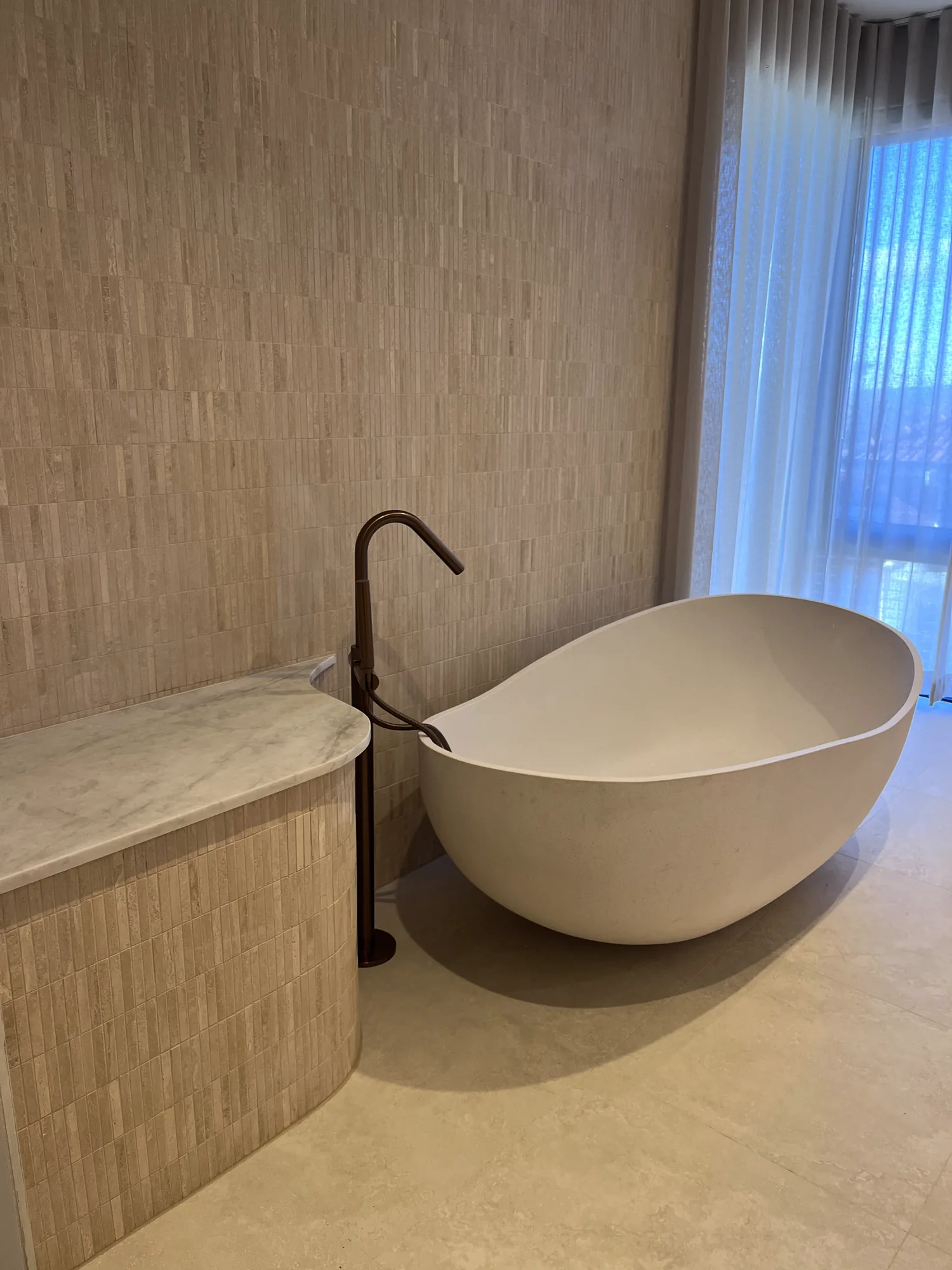 Freestanding bath beside curved tiled joinery and a bronze floor-mounted tap.